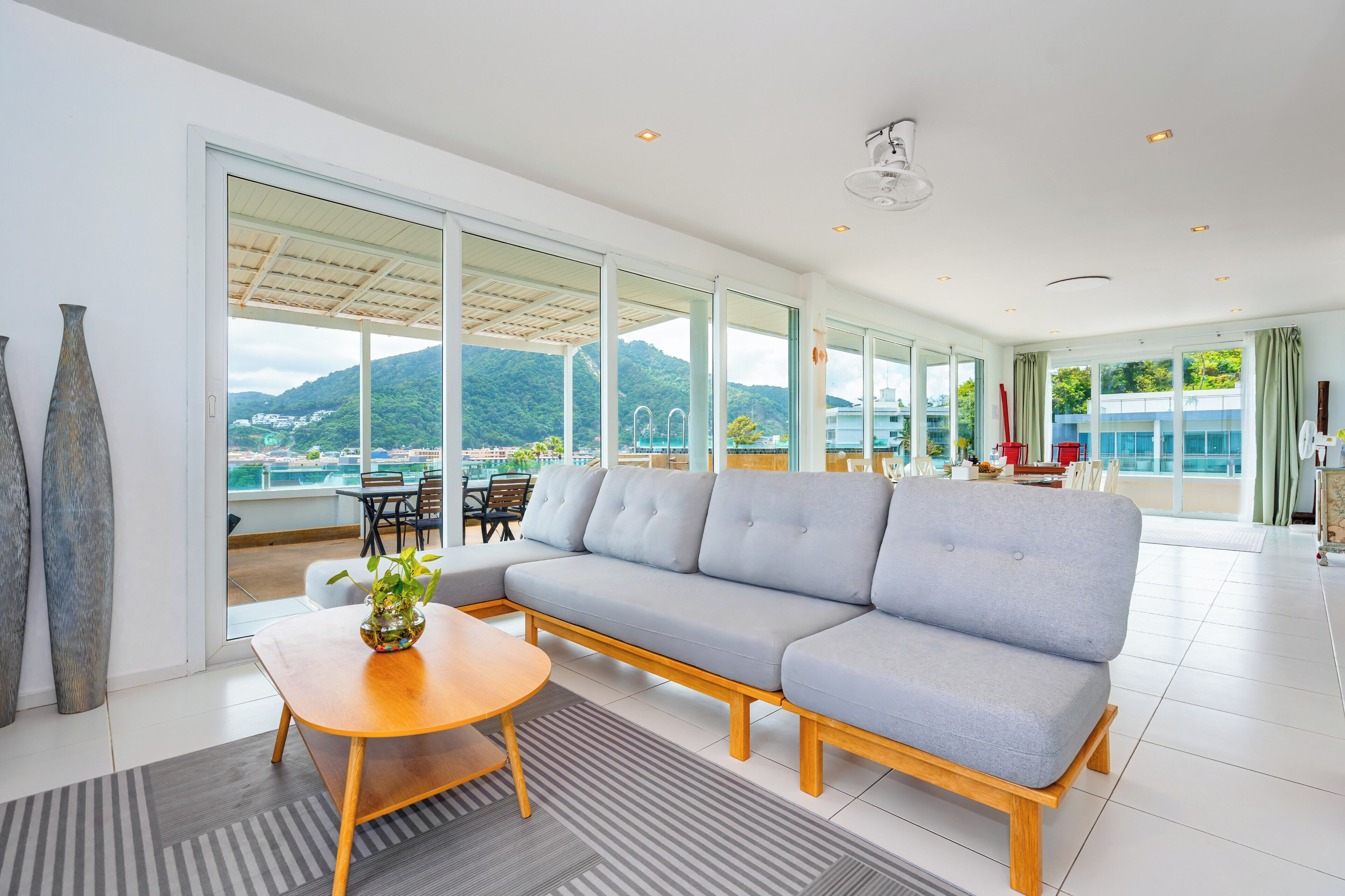 Una sala de estar moderna con un sofá gris, una mesa de centro de madera y ventanas amplias que ofrecen vistas a un paisaje montañoso.