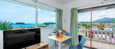A modern room with a TV, dining table, chairs, and a balcony with a view of the sea and mountains.