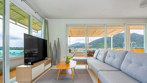 A modern living room with a large flat-screen TV, a wooden coffee table, a grey sofa, and a view of a mountainous landscape through large windows.