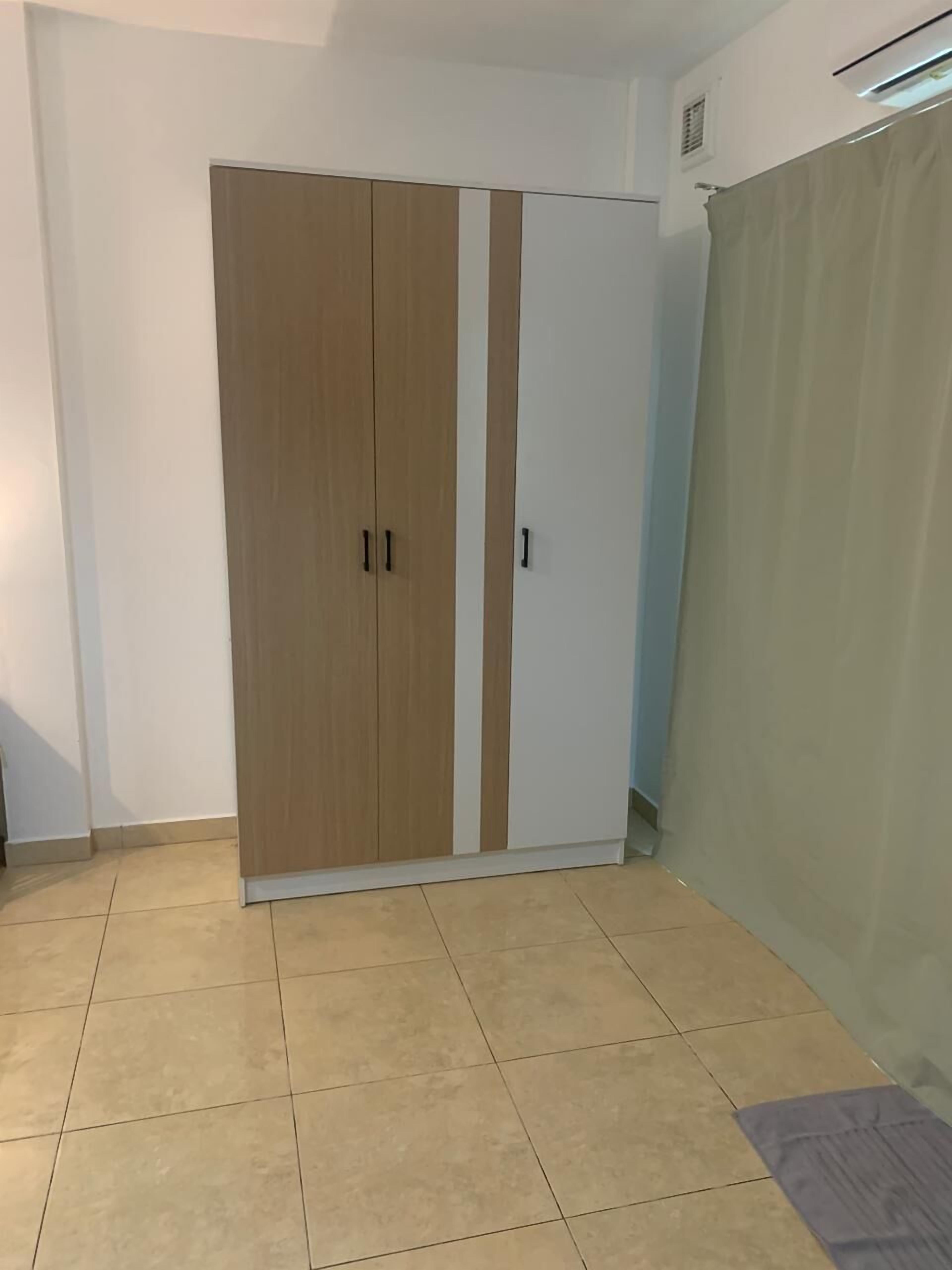 Habitación con armario de dos puertas, una con detalles de madera y la otra blanca. El piso es de baldosas y hay una cortina a la derecha.
