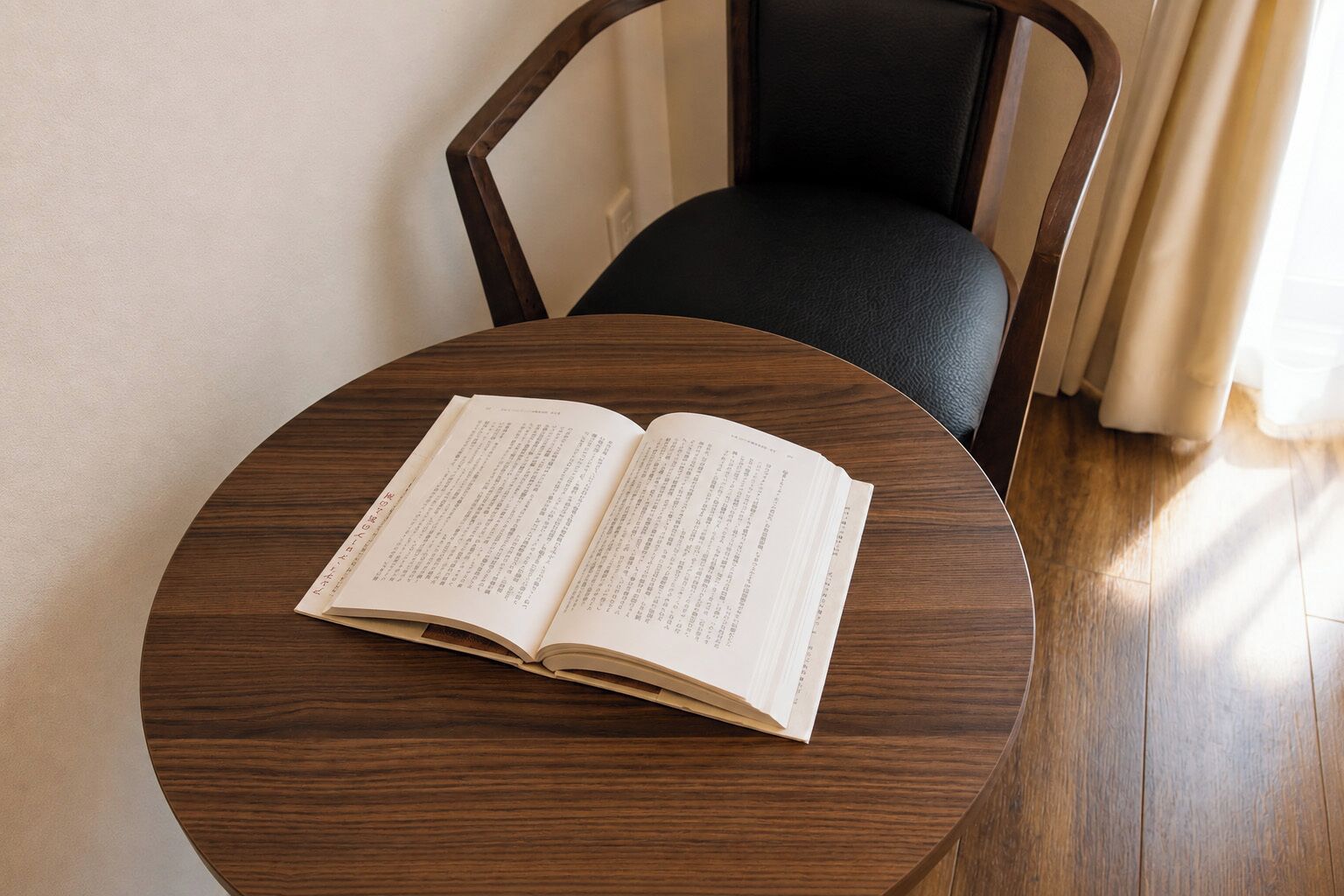 A round wooden table with an open book on it, positioned in front of a chair with a black cushion.