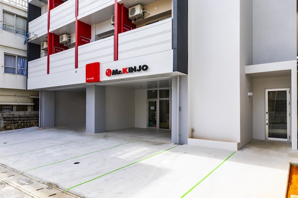 Un edificio moderno con un llamativo letrero que dice 'Mr. Kinjo', un área al aire libre techada con líneas delimitadas y una escalera que conduce a un nivel superior.