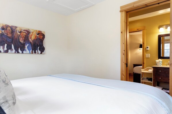 A bedroom with a bed, a painting of bulls on the wall, a wooden door, and a bathroom visible through an open door. - Outpost: Alpine Cottage Two (Jackson)