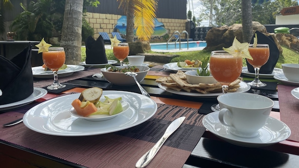 A table set with breakfast items including fruit, bread, and drinks, with a pool and palm trees in the background. - Rohoney Boutique Villa (Galle)