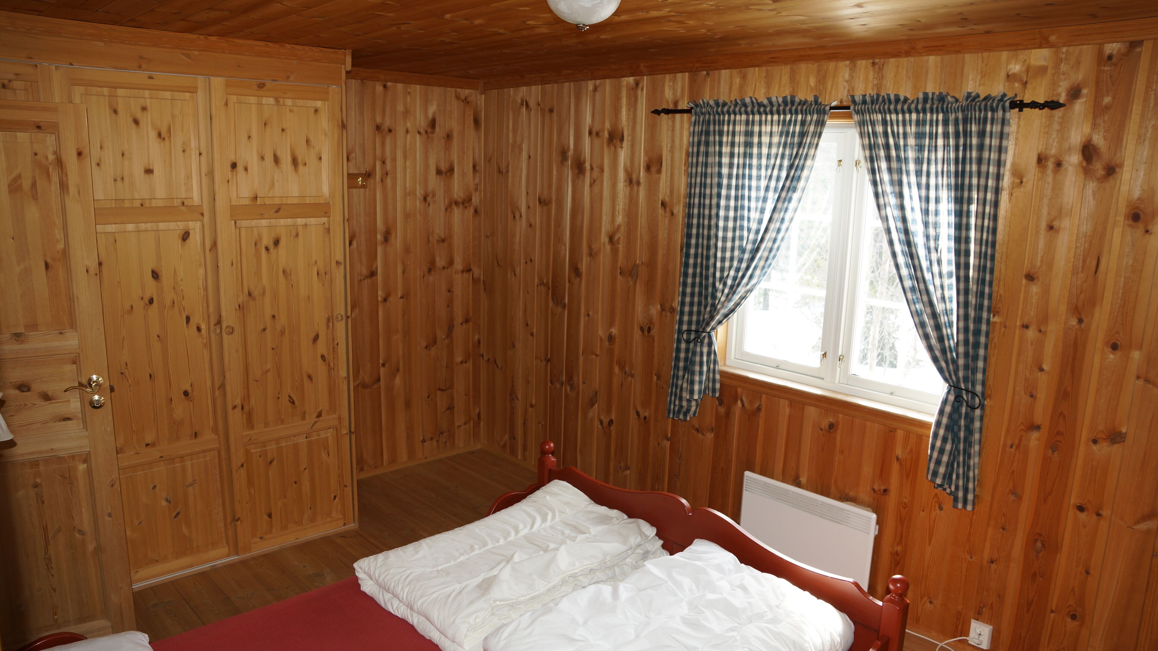 A wooden-paneled room with a bed, a window with curtains, and a radiator.