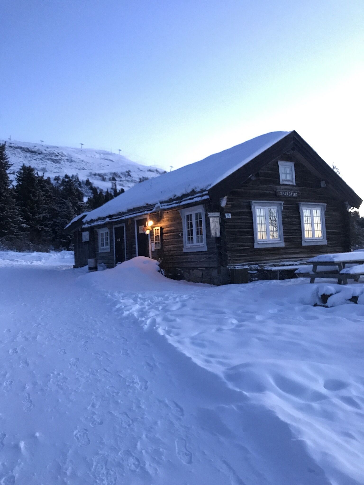 A wooden cabin with a snow-covered roof and a sign reading "SkiStua."