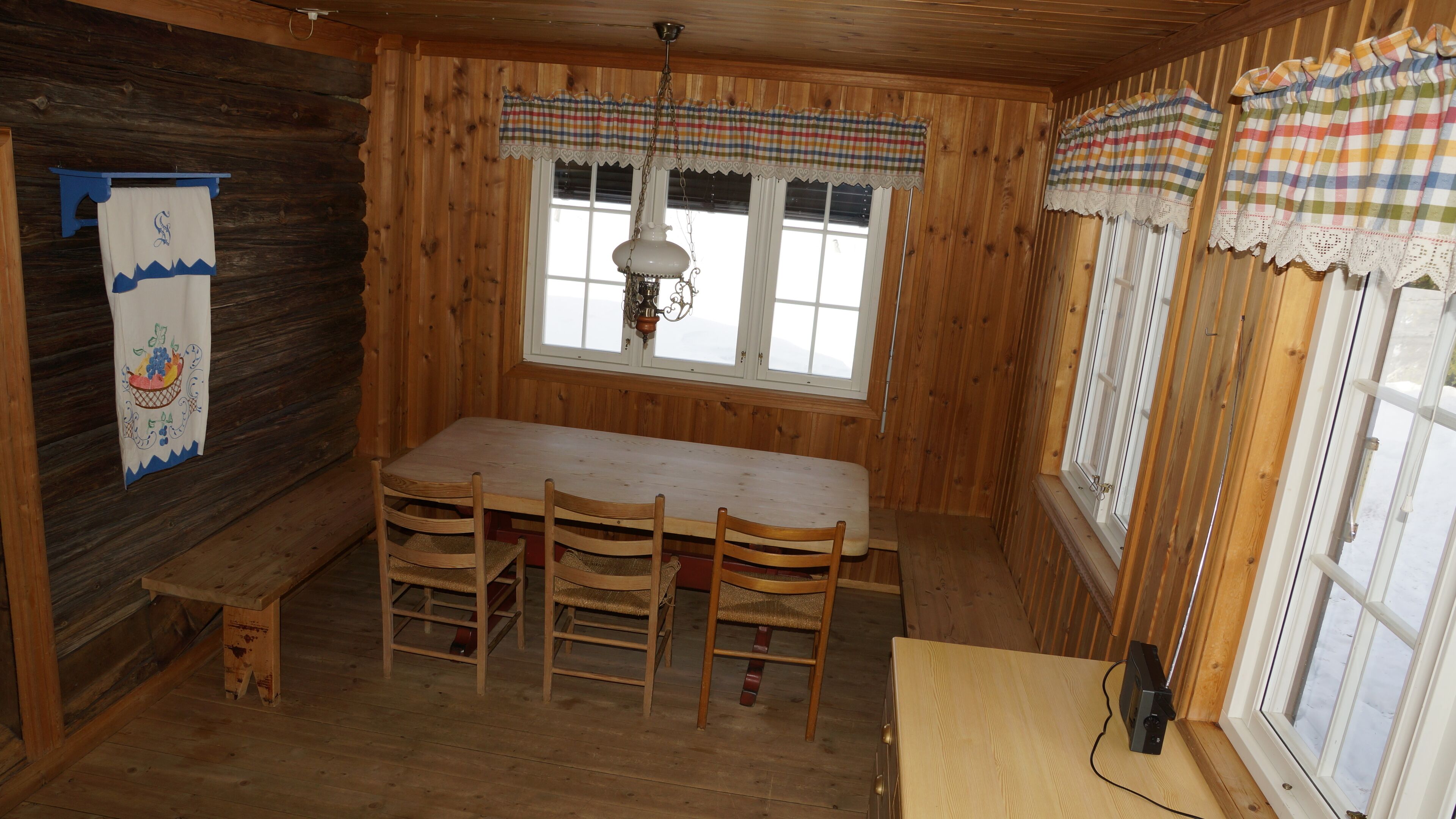 A wooden cabin interior with a dining table, four chairs, a hanging light fixture, and a window with plaid curtains.
