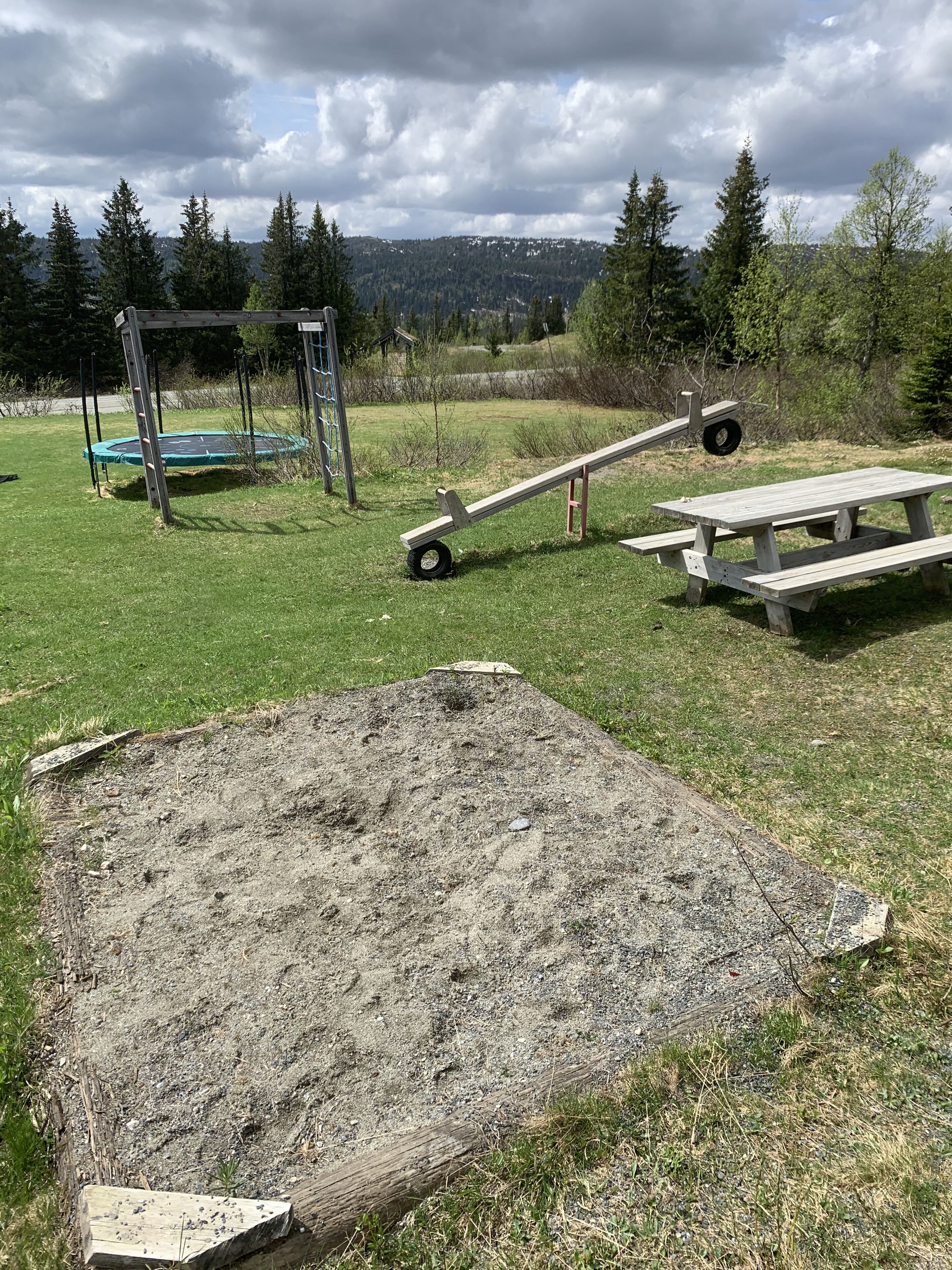 A playground with a swing set, a seesaw, and a picnic table in a grassy area with trees in the background.