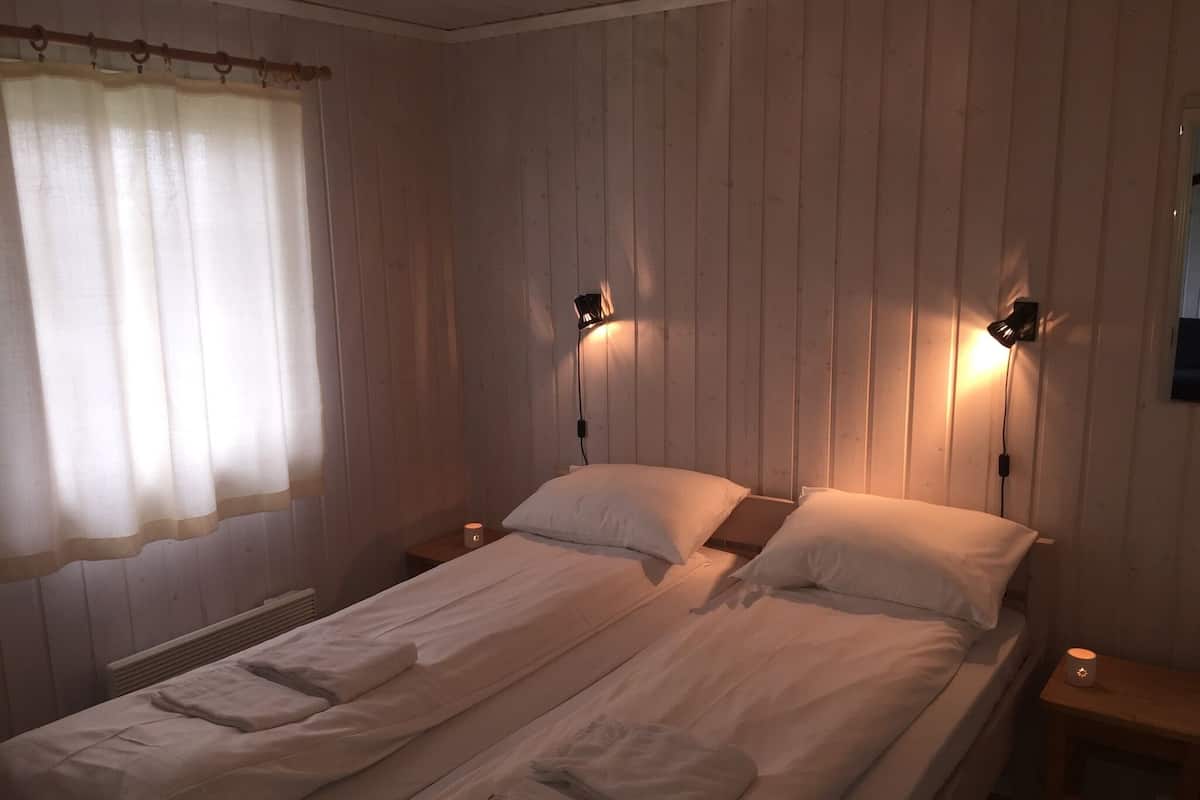 Una cama doble con ropa de cama blanca, dos mesitas de noche con lámparas y un ventanal con cortina ligera.