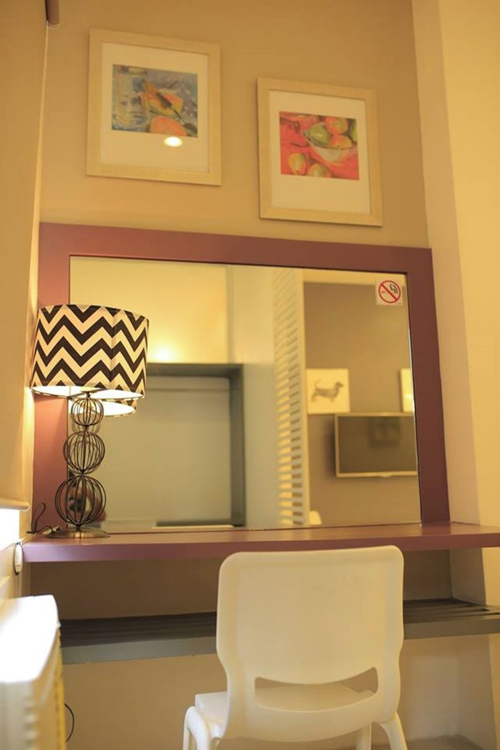 A hotel room with a large mirror, a white chair, a lamp with a chevron pattern, and two framed artworks on the wall.