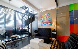 Um saguão de hotel moderno com um sofá de couro preto, um banquinho branco, uma poltrona preta, uma mesa redonda e uma mesa de centro de vidro. Há uma pintura abstrata colorida na parede, uma escada em espiral e uma grande janela com persianas.