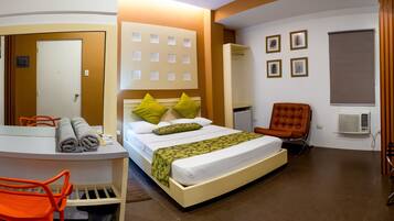 Habitación de hotel con cama, escritorio y silla, espejo, radiador y ventana con persianas.