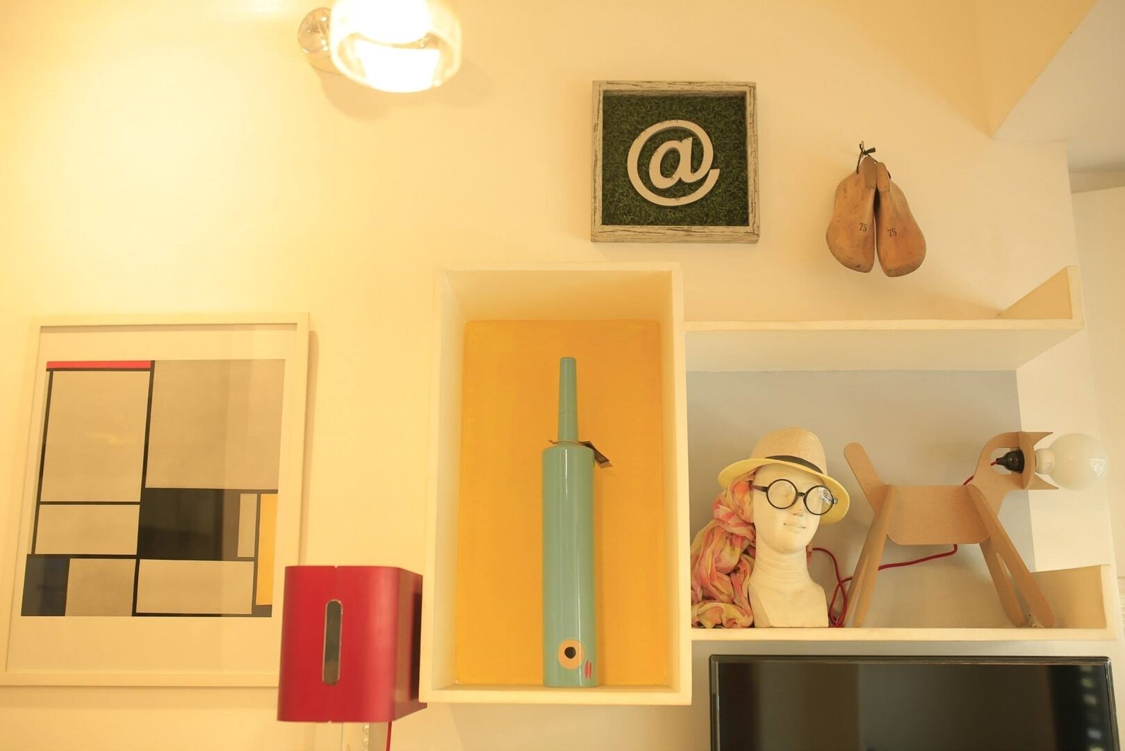 A room with a framed abstract painting, a wooden sculpture, a mannequin with a hat, and a light fixture.