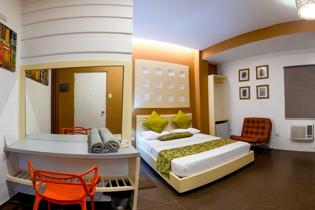 A hotel room with a desk, a bed with green and white bedding, a chair, and a TV.