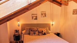 A cozy bedroom with a sloped ceiling, a bed with a white quilt, and two deer-themed pillows. There are wall-mounted lights and framed pictures above the bed. - Lezegorria (Urzainqui)