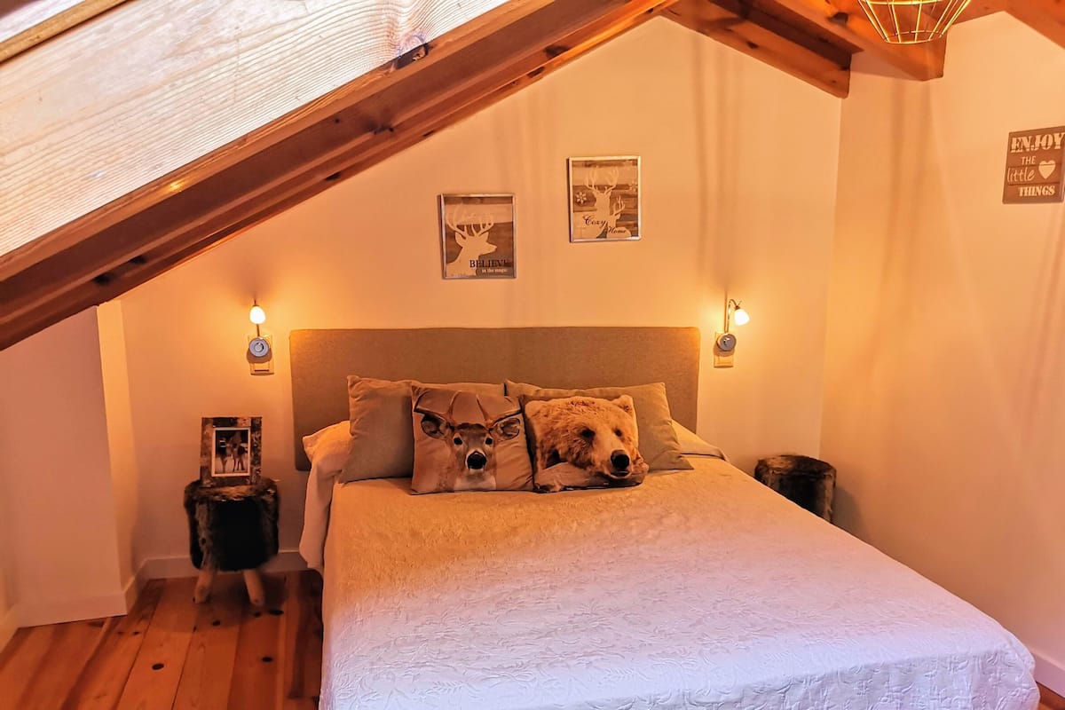Ein gemütliches Schlafzimmer mit schrägen Wänden, einem Bett mit weißer Bettdecke und zwei Kissen mit Hirschmotiven. An den Wänden sind Wandleuchten und Bilder in Rahmen angebracht.