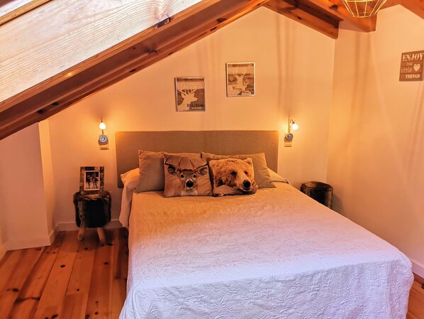 Een gezellige slaapkamer met een schuine kap, een bed met een witte quilt en twee kussens met hertenmotieven. Er hangen wandlampen en lijstwerken boven het bed.