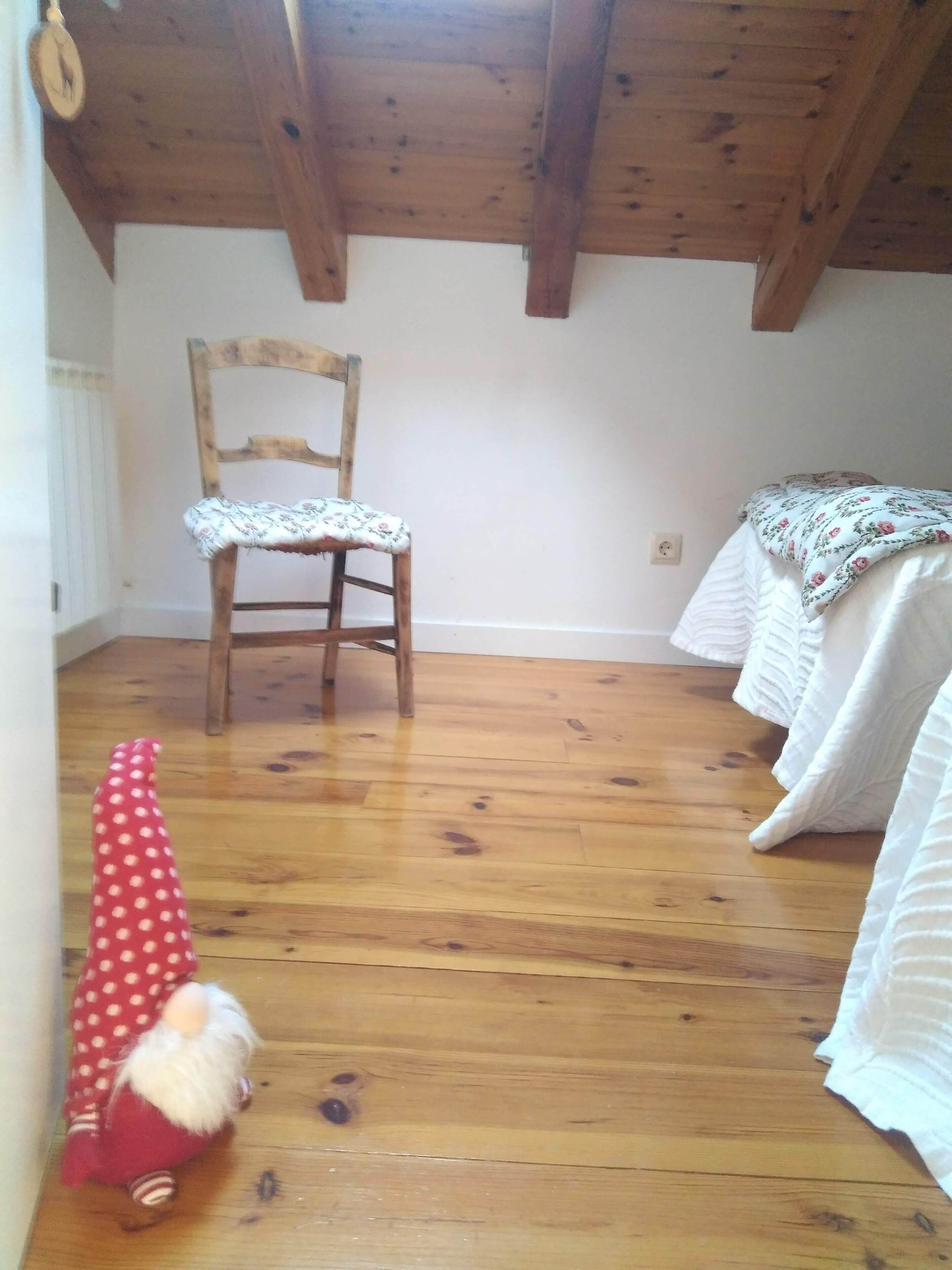 A room with a wooden floor, a wooden chair, a white bed with a floral bedspread, and a red polka-dotted object on the floor.