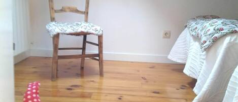 A room with a wooden floor, a wooden chair, a white bed with a floral bedspread, and a red polka-dotted object on the floor.