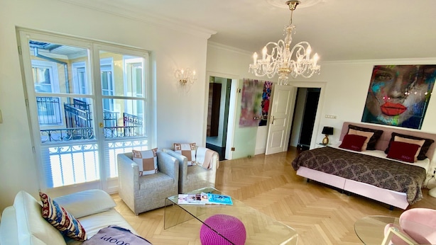 Un salon spacieux avec un canapé blanc, une table basse en verre et un pouf rose. On y trouve un lit avec une literie à motifs et un grand tableau accroché au mur.