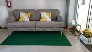 A grey sofa with two yellow and white patterned cushions, a green rug, and a small wooden side table with a vase of flowers. - Prinya House (Chonburi)