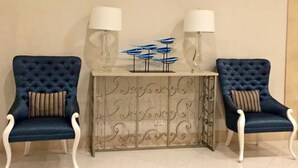 Una habitación con dos sillones tapizados en azul con reposabrazos blancos, una mesa central con cubierta de cristal decorativa y dos lámparas fijadas a la pared.