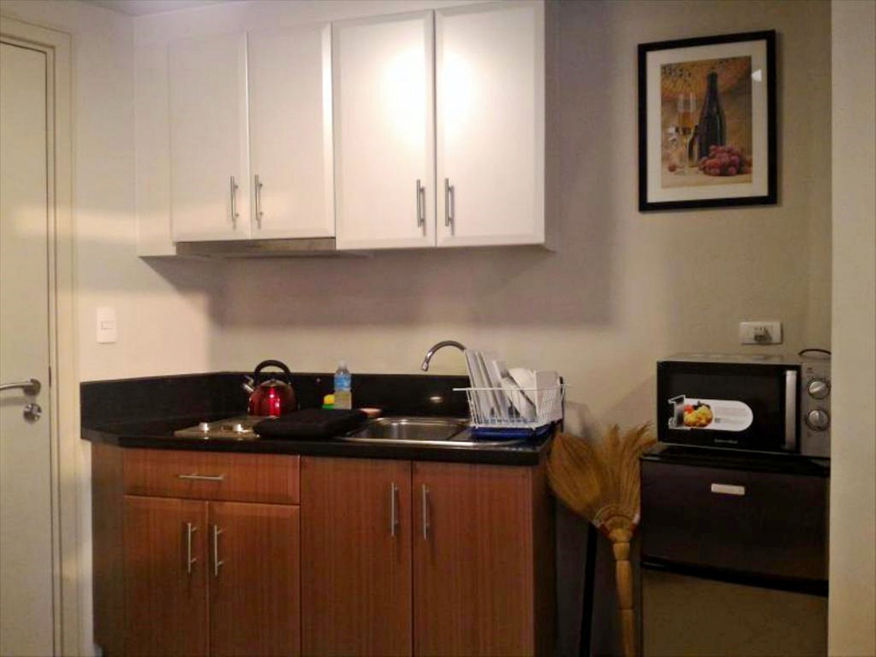 A compact kitchen with wooden cabinets, a kettle, a sink, a broom, and a microwave.