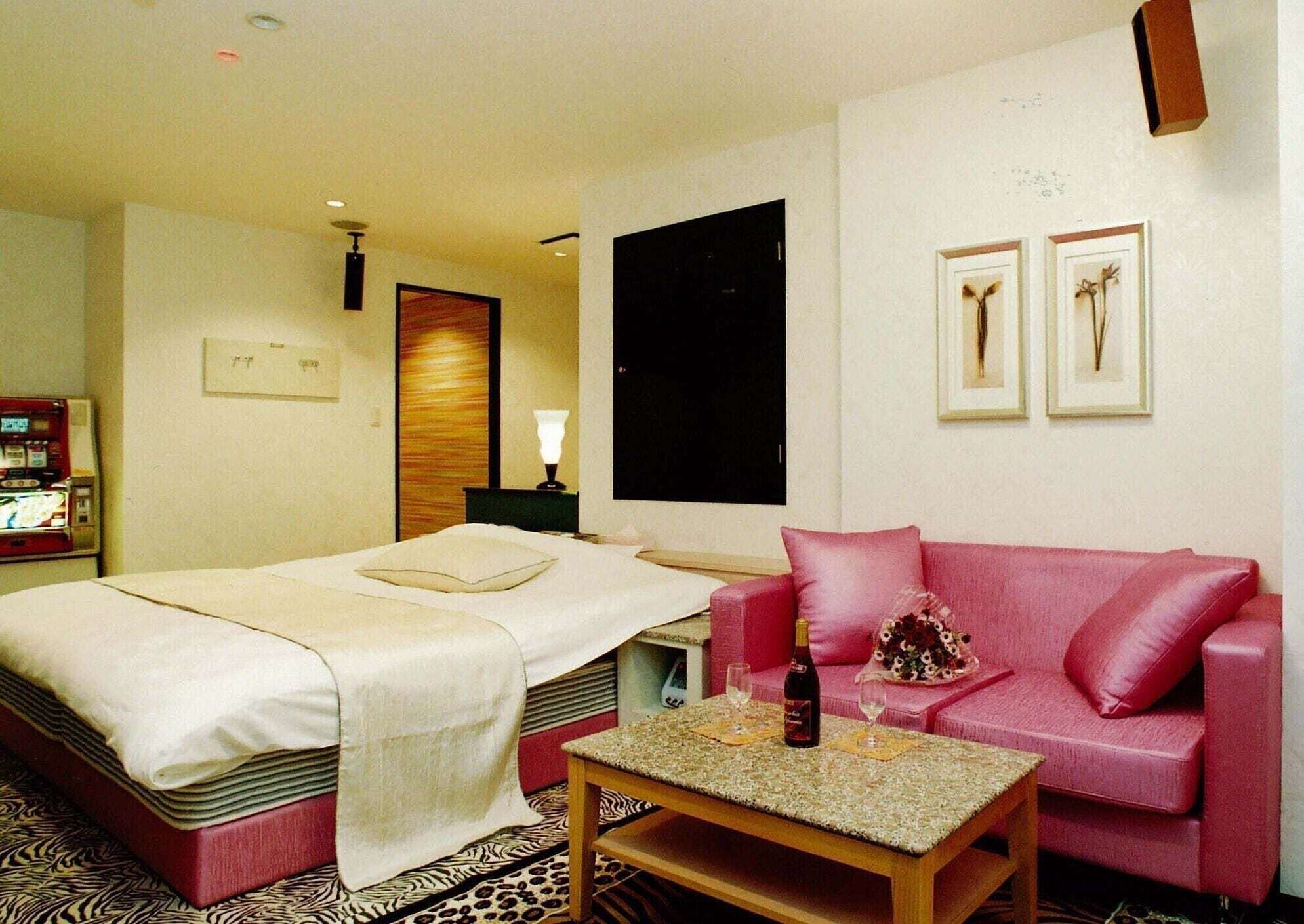 Quarto de hotel com cama, sofá vermelho, mesa pequena com uma garrafa e copos, e televisão fixada na parede.