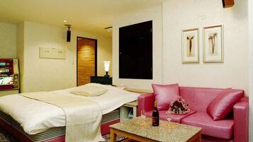 Quarto de hotel com cama, sofá vermelho, mesa pequena com uma garrafa e copos, e televisão fixada na parede.