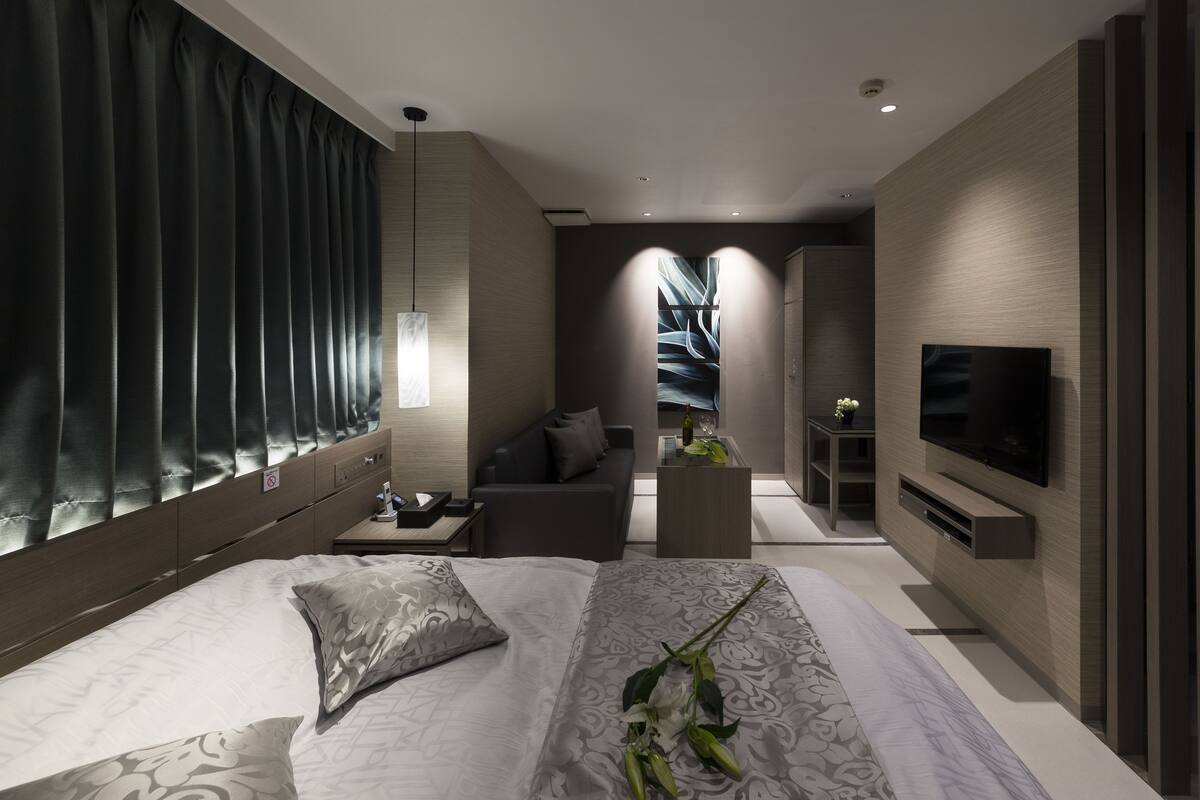 Une chambre d’hôtel moderne avec un grand lit, un canapé, une télévision et une petite table sur laquelle se trouvent une bouteille et des verres.