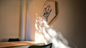 A dining area with a wooden table and chairs, a framed artwork on the wall, and sunlight casting shadows. - Yes Yes I am Willing (Tainan)