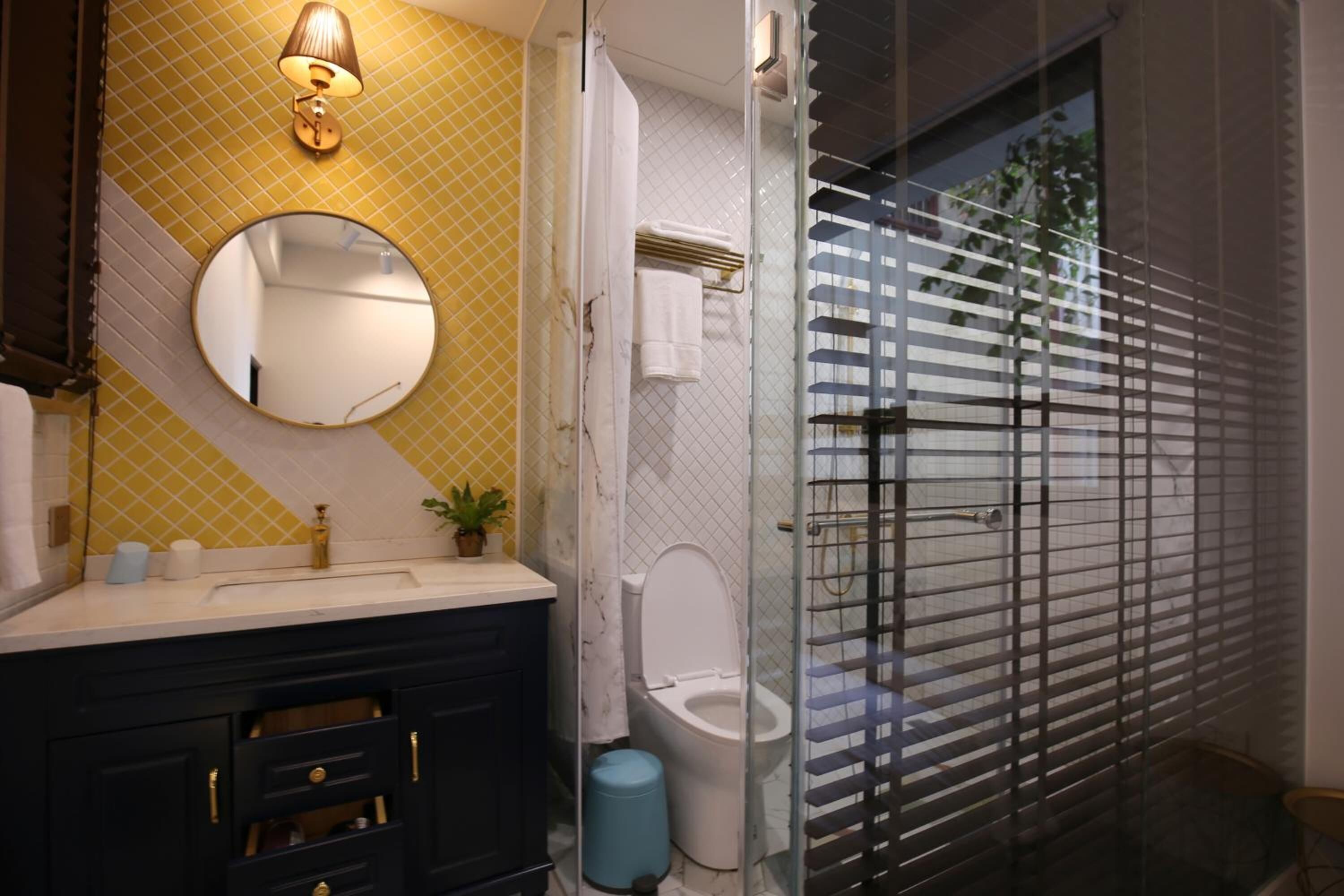 A bathroom with a yellow patterned wall, a round mirror, a sink with a gold faucet, a toilet, and a glass shower enclosure with a window.