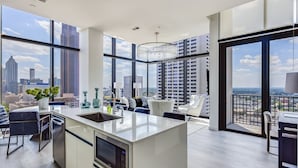 A modern kitchen with a large island, white cabinetry, and a view of the city skyline through large windows. - Vue Spectaculaire Penthouse (Atlanta)