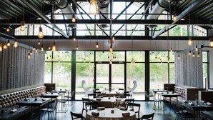 A modern restaurant with high ceilings, exposed beams, and large windows offering a view of greenery. Tables are arranged for dining, and there are cushioned seating areas. - Westside Retreat (Atlanta)