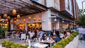 Un'area esterna con tavoli e sedie per mangiare, l'ingresso di un ristorante e la facciata di un edificio con un tetto sporgente in legno e luci a sospensione.