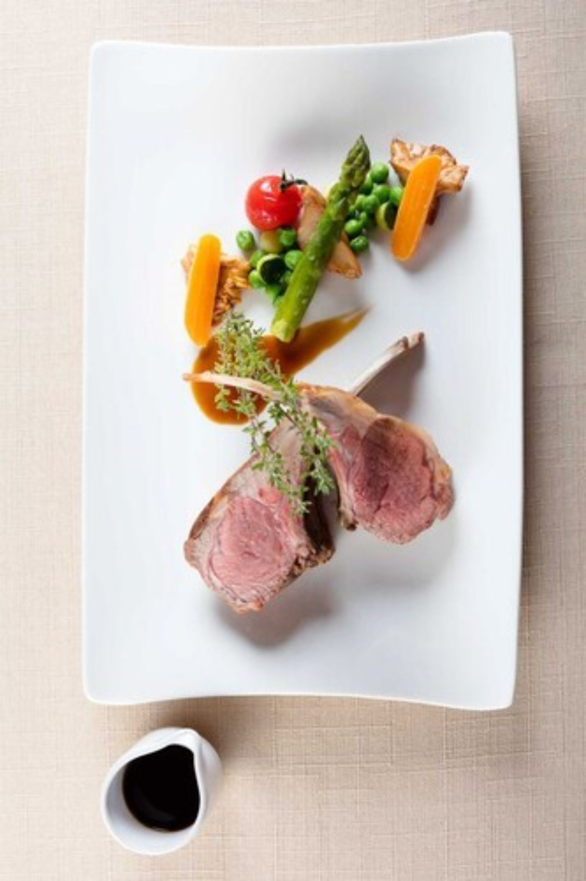Une assiette blanche contenant un morceau de viande, des asperges, des petits pois et une tomate cerise.