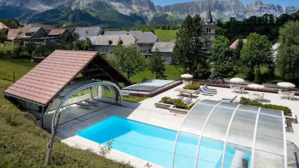 Un espace piscine abrité par une structure en verre, entouré de maisons et de montagnes.