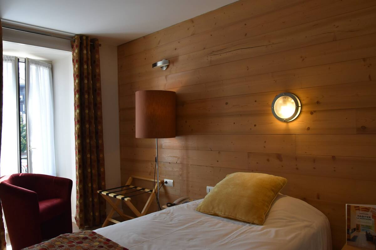 Une chambre d’hôtel avec un mur en bois, un lit avec un oreiller jaune, une chaise rouge, une lampe et une fenêtre avec des rideaux.