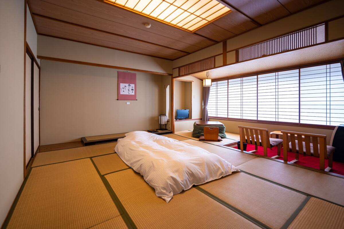 A traditional Japanese room with tatami flooring, a futon bed, a television, and a sliding door leading to a balcony.