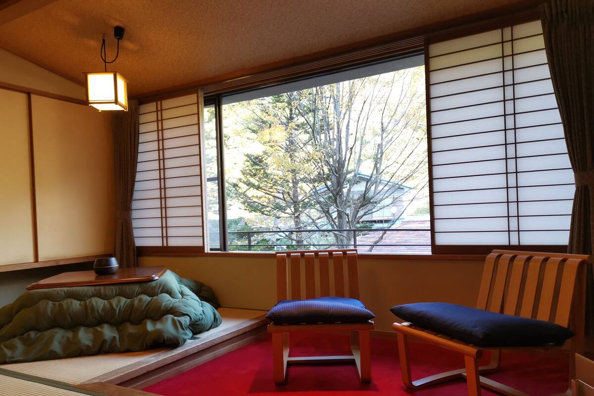 A traditional Japanese room with tatami flooring, a low table with a cushion, two wooden chairs, and a window with shoji screens.