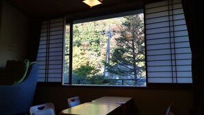 A room with tatami mat flooring, a low table, and chairs. A large window with shoji screens provides a view of trees and a building outside.