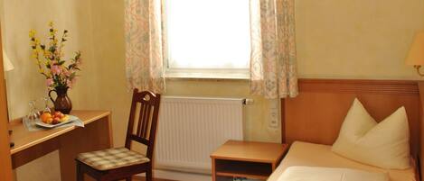 A hotel room with a bed, a desk, a chair, a window with curtains, and a radiator.