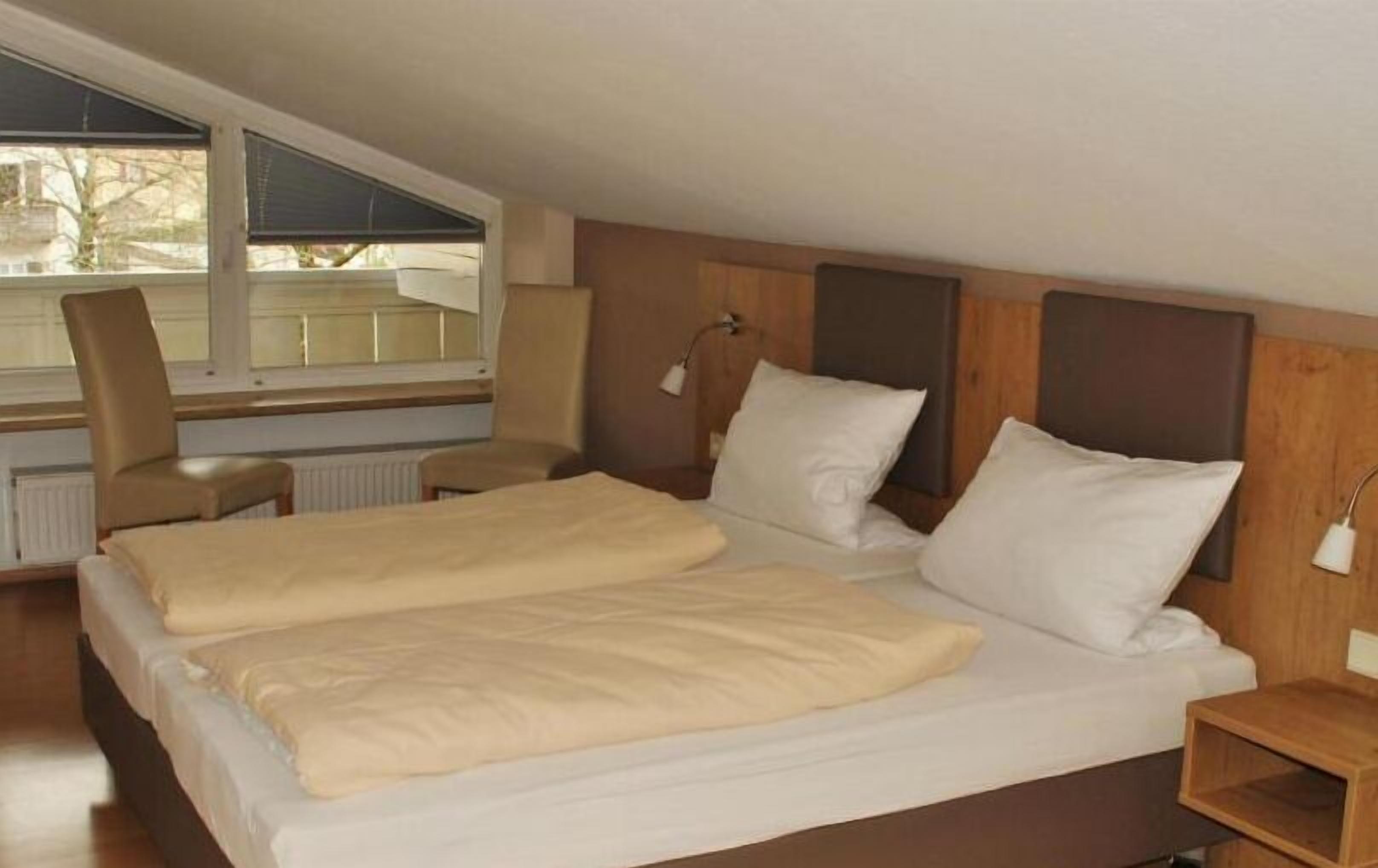 A twin bed with a beige blanket, a wooden headboard, and a window with blinds.