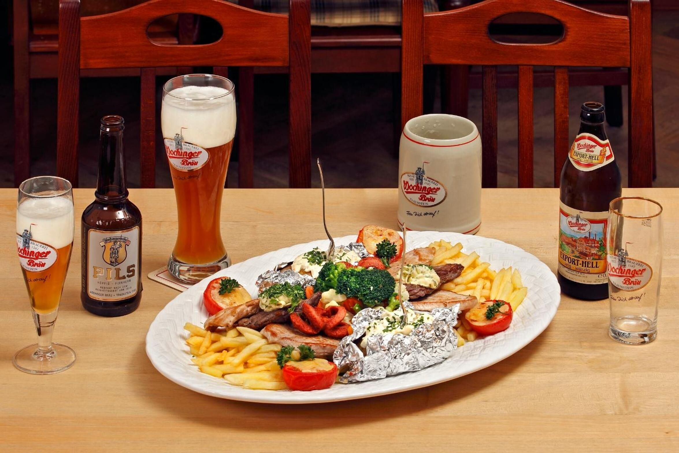 Una mesa de comedor con un plato de comida, varios vasos y botellas de cerveza, y sillas de madera.