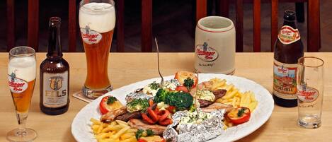 Una mesa de comedor con un plato de comida, varios vasos y botellas de cerveza, y sillas de madera.