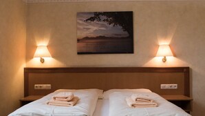 Un lit double avec deux tables de chevet, chacune équipée d’une lampe et d’une prise électrique. Un tableau représentant un paysage est accroché au-dessus du lit.