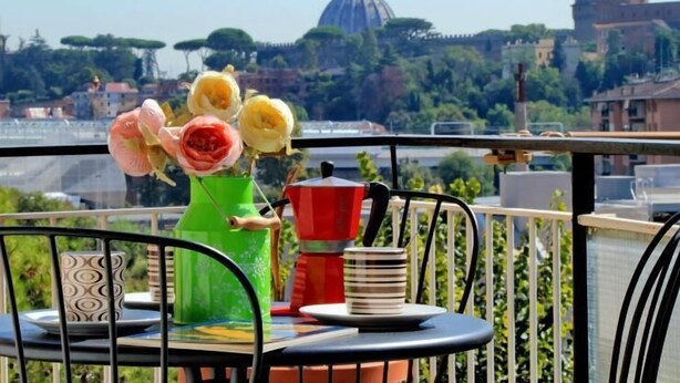 Un balcón con una mesa, sillas, un jarrón con flores, una cafetera y vistas a una cúpula y árboles.