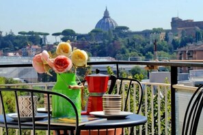 Uma varanda com mesa, cadeiras, um vaso de flores, uma cafeteira e vista para uma cúpula e árvores.