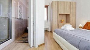 Una habitación de hotel moderna con una cama grande, un armario de madera, ducha de vidrio y una pequeña zona de estar.