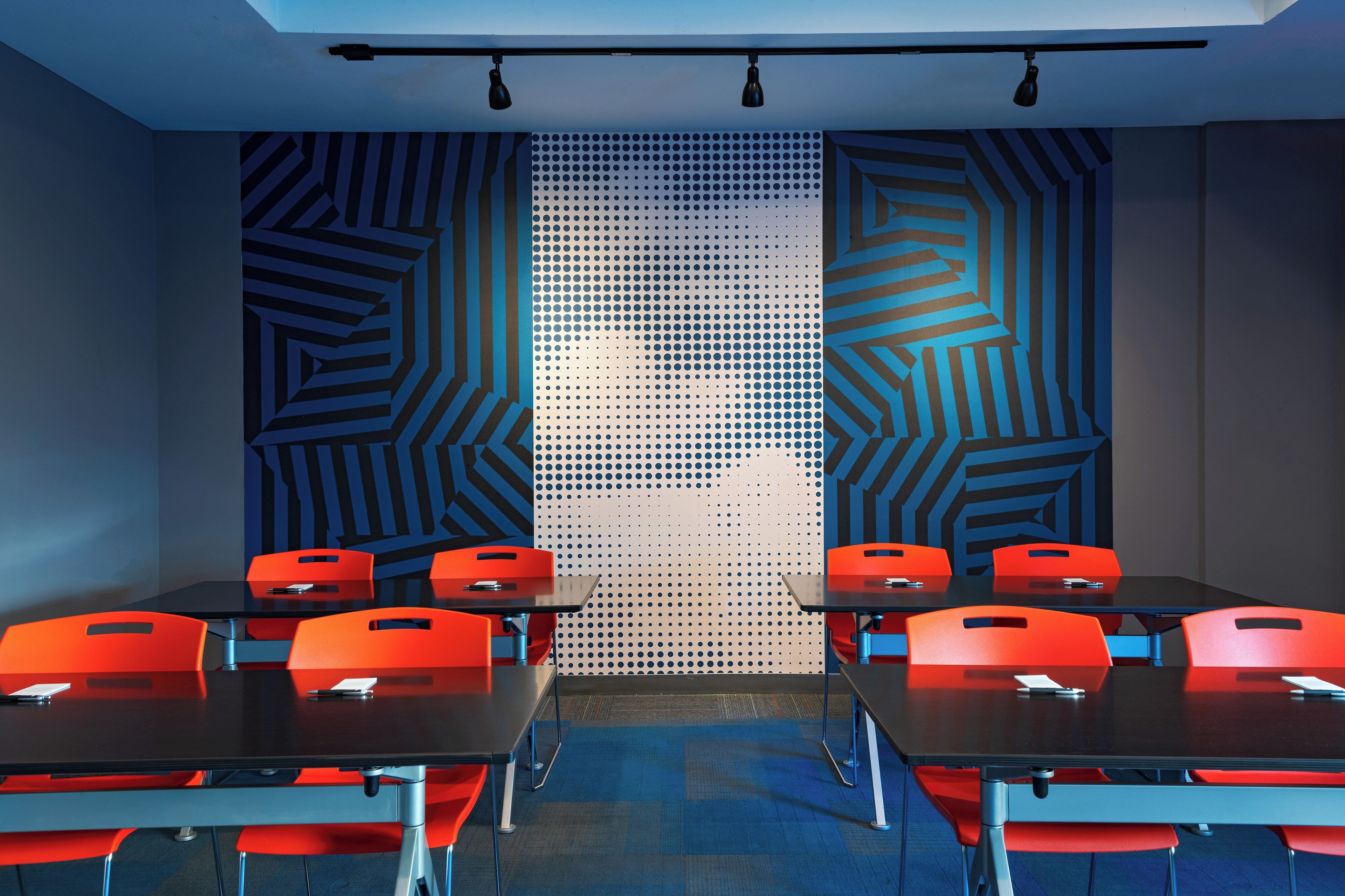 Un salón de conferencias con diseño moderno, que presenta una pared con estampado geométrico, un panel perforado y sillas rojas alrededor de una mesa central.